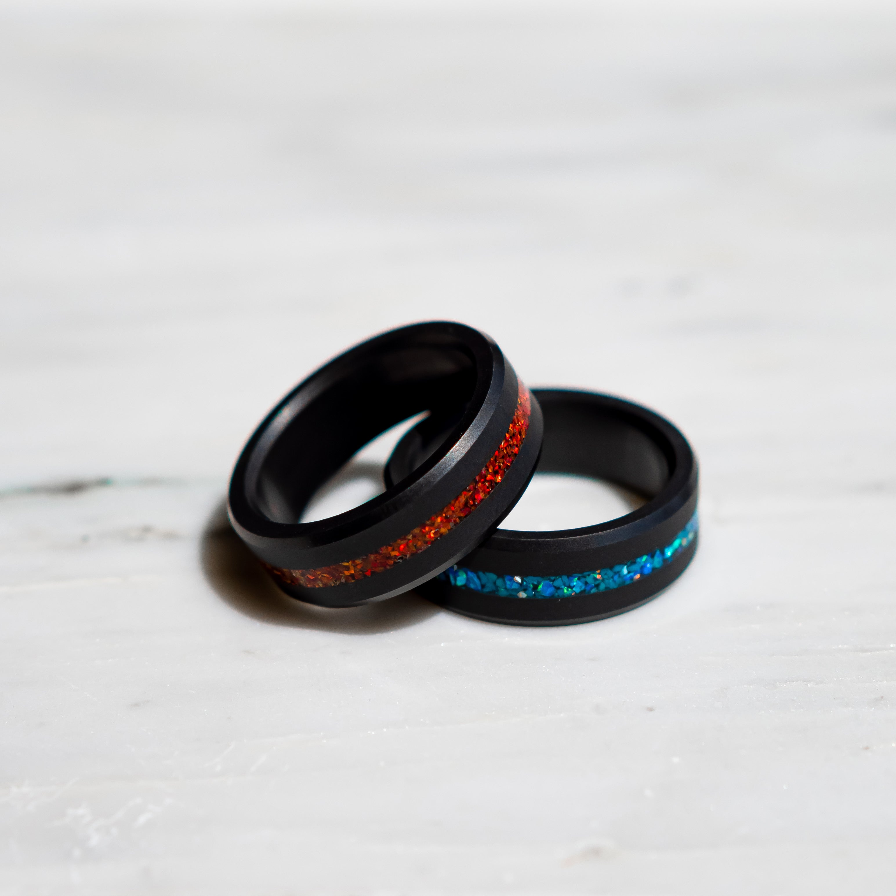 Two solid black diamond rings with red and blue opal inlay on a white marble surface