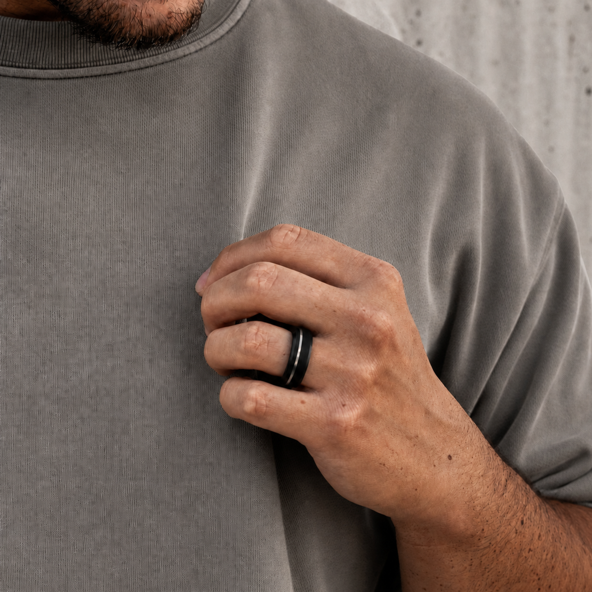 A close-up of a man's hand wearing a a Solid Black Diamond wedding band with a silver offset inlay. Shown against his gray sweatshirt and a concrete wall background.