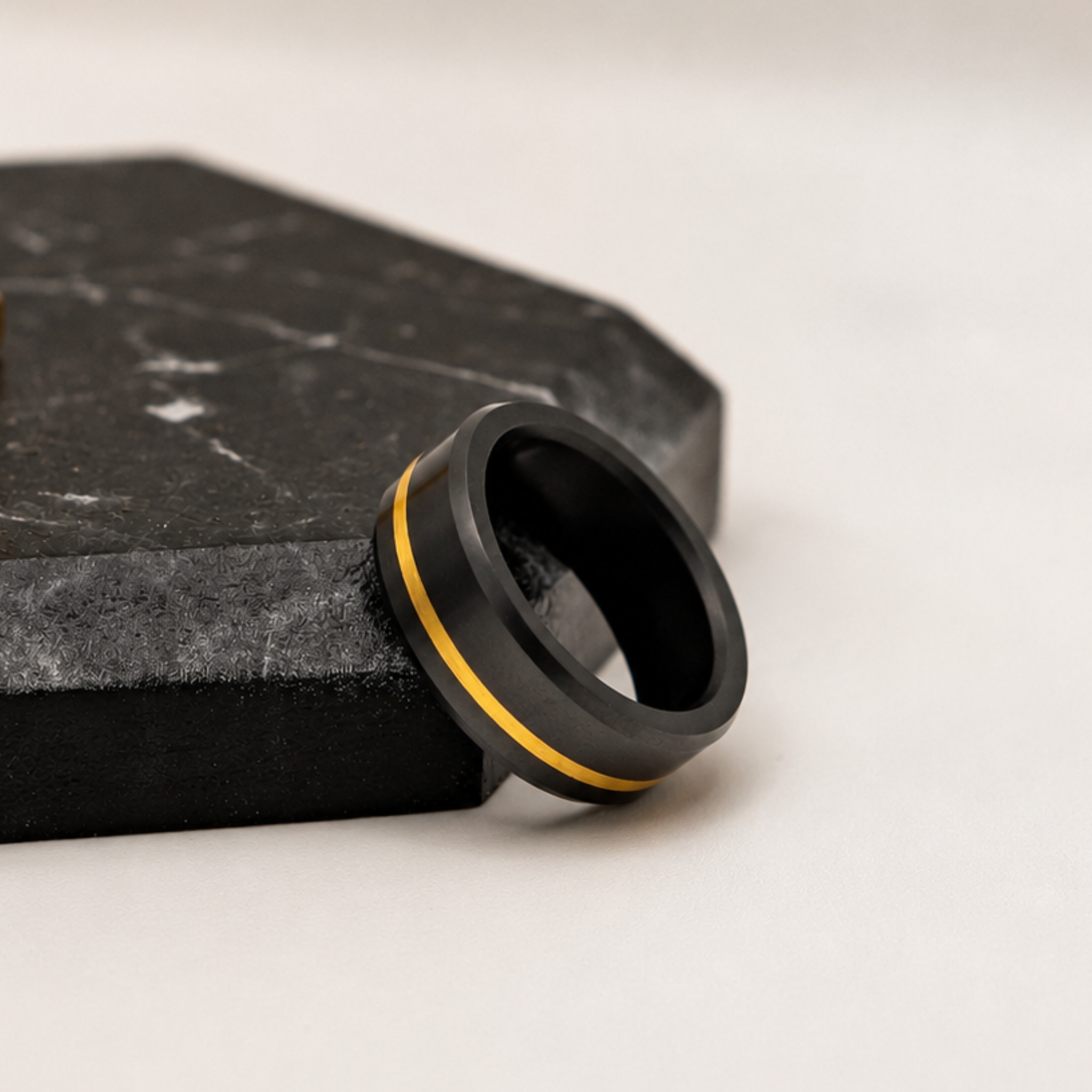 solid black diamond ring with a 24K yellow gold offset inlay, resting on a dark black stone slab against a light off-white background